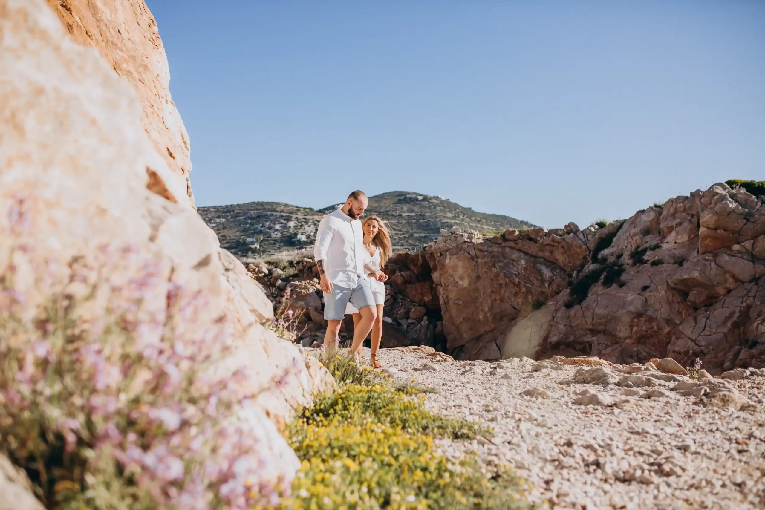 Pareja caminando por un sendero de tierra y rocas entre formaciones rocosas en un día soleado, ideal para excursiones faciles mallorca. Flores en primer plano.